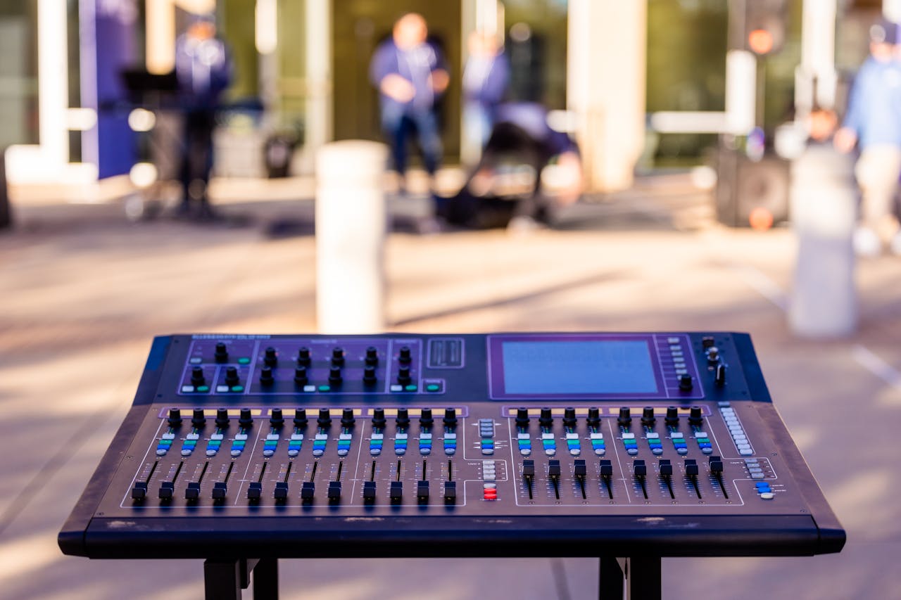 Focus on a digital mixing console outdoors with blurred musicians in the background, capturing a live music scene.